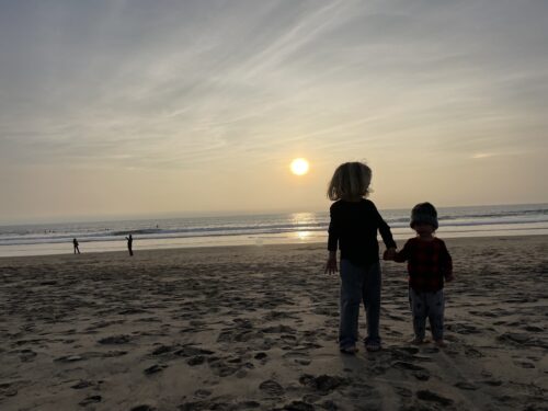 To young children hold hands on a beach close to sunset. In the foreground, sand is covered in footsteps. The ocean is calm beneath a setting sun.