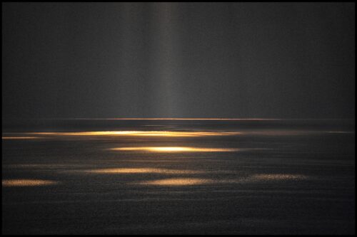 A darkened image of the sky and ocean with pillars of light streaming down, creating golden reflections on the water. 